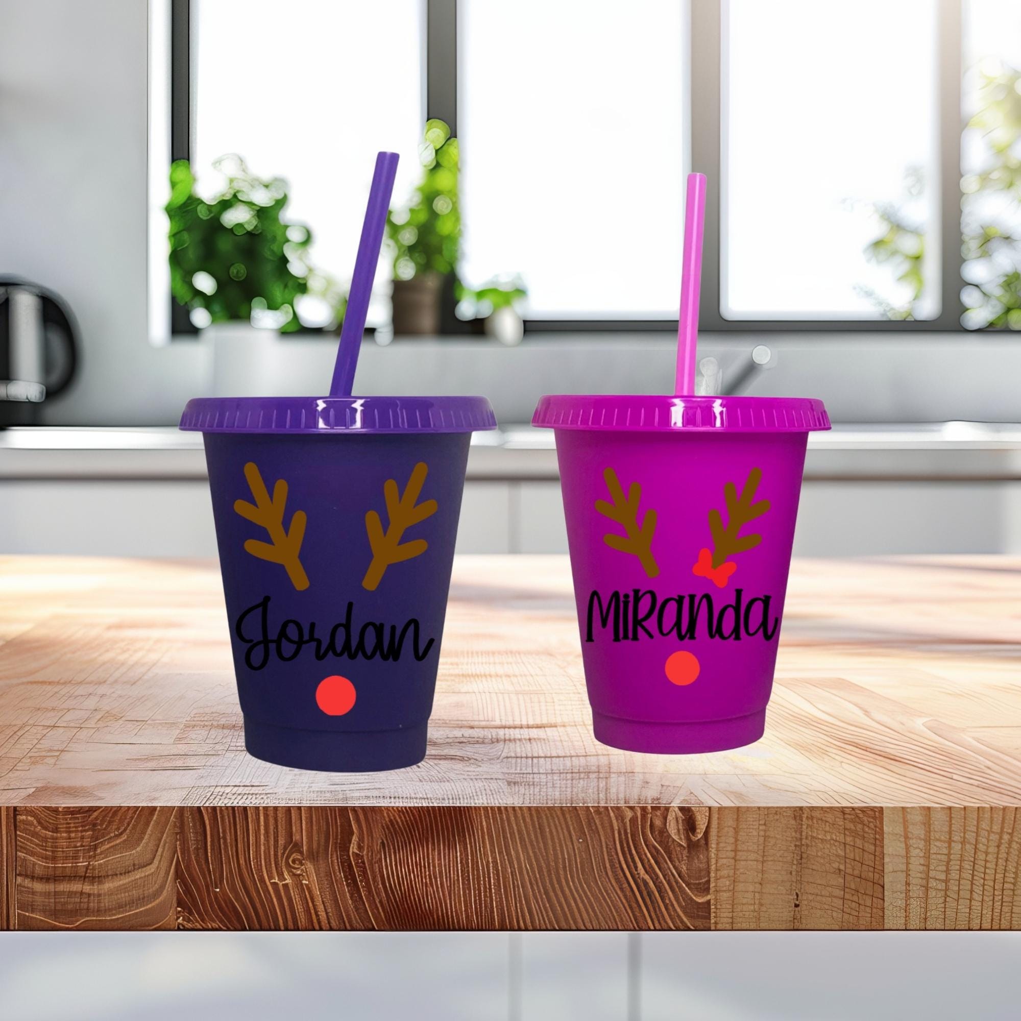 Two personalized cups with reindeer designs and names on a wooden surface.